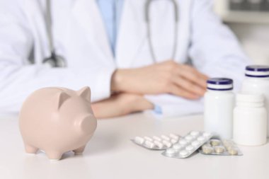 Doctor at white table indoors, focus on piggy bank and pills