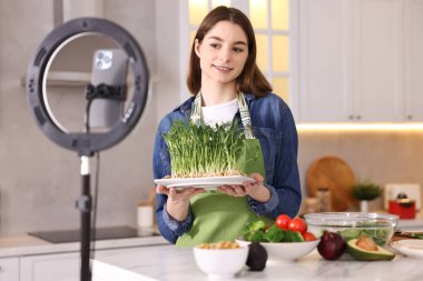 Yemek blogcusu mutfakta akıllı telefon ve yüzük lambasıyla video kaydederken yemek pişiriyor.