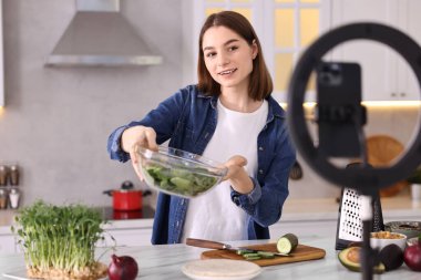 Yemek blogcusu mutfakta akıllı telefon ve yüzük lambasıyla video kaydederken yemek pişiriyor.