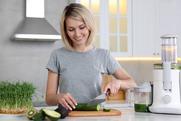 Juicer and fresh products on white marble table. Smiling woman cutting cucumber in kitchen