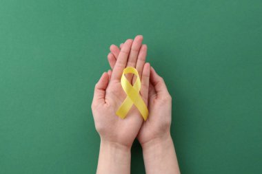 Woman with yellow awareness ribbon on green background, top view