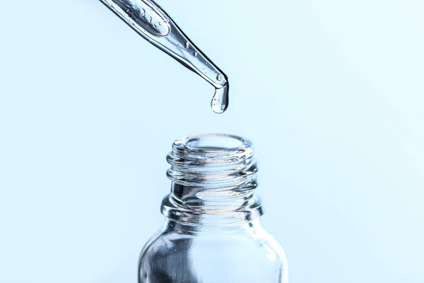 Dripping liquid from pipette into glass bottle on light blue background, closeup