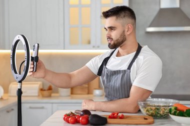 Yemek blogcusu mutfakta akıllı telefon ve yüzük lambasıyla video kaydederken yemek pişiriyor.