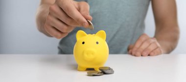 Beyaz masadaki sarı kumbaraya para koyan adam, yakın plan.