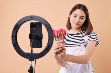 Güzellik blogcusu makyaj fırçalarını inceliyor ve arka planda akıllı telefon ve yüzük lambasıyla video kaydediyor.