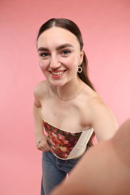 Şık korseli, gülümseyen kadın pembe arka planda selfie çekiyor.