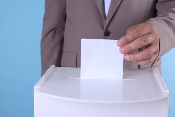 Man putting his vote into ballot box against light blue background, closeup