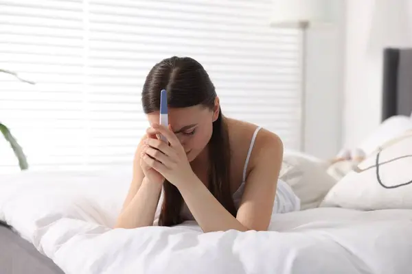 stock image Sad woman holding pregnancy test on bed in room