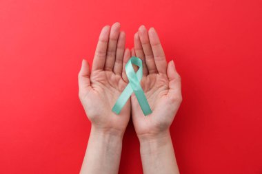 Woman holding turquoise awareness ribbon on red background, top view clipart