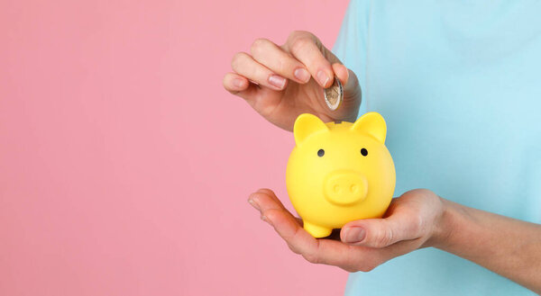 Woman putting coin into yellow piggy bank on pink background, closeup. Space for text