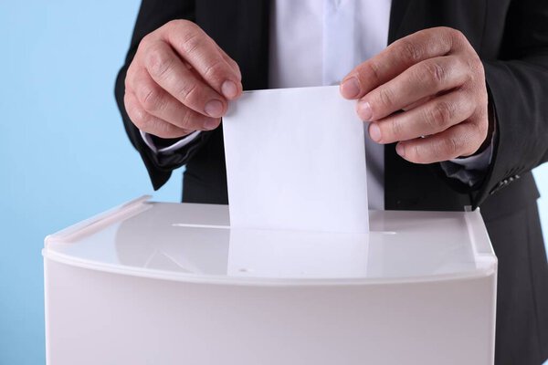 Man putting his vote into ballot box against light blue background, closeup