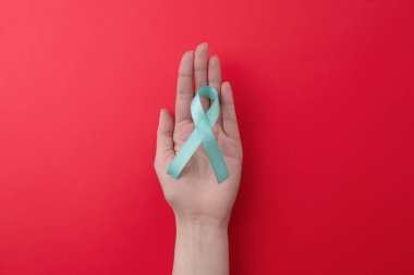 Woman holding turquoise awareness ribbon on red background, top view