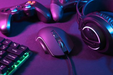 Bilgisayar faresi, kulaklık, RGB klavye ve oyun denetleyicileri koyu arkaplanda neon ışıkları, yakın plan