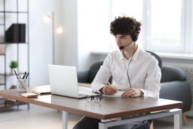 İçerideki masada dizüstü bilgisayarla çalışırken not alan kulaklıklı bir genç. Uzak görev