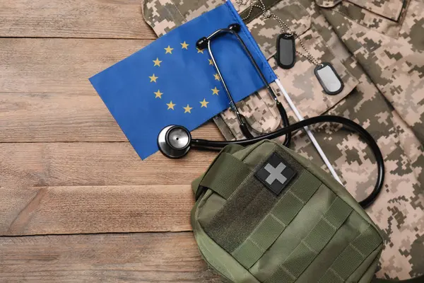 stock image Stethoscope, flag of European Union, first aid kit and military uniform on wooden table, flat lay. Space for text