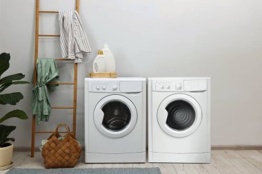 Çamaşır odasındaki açık gri duvarın yanında iki çamaşır makinesi.