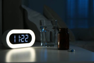 Insomnia treatment. Glass of water, pills and alarm clock on bedside table in bedroom at night clipart