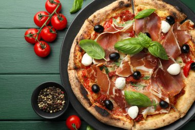 Tasty pizza with cured ham, olives, mozzarella cheese, tomatoes and arugula on green wooden table, flat lay