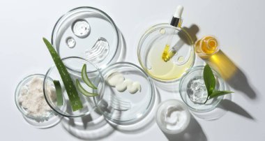 Beyaz arka planda farklı kozmetik ürünleri ve aloe vera yaprakları içeren petri kapları, düz yatıyordu