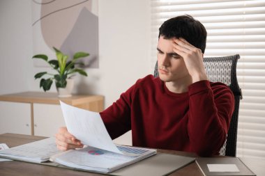 Embarrassed man with document at table in office clipart