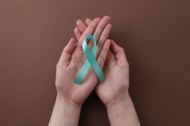 Woman holding turquoise awareness ribbon on brown background, top view clipart