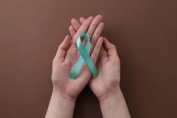 stock image Woman holding turquoise awareness ribbon on brown background, top view