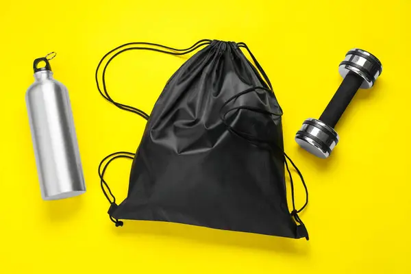 stock image Black drawstring bag, thermo bottle and dumbbell on yellow background, flat lay