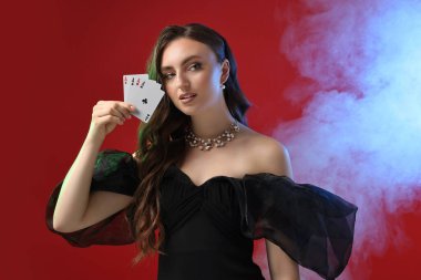 Poker oyunu. Kırmızı arka planda iskambil kağıtlarıyla renkli ışıklar ve dumanla oynayan büyüleyici bir kadın.