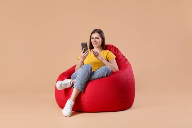 Bej arka planda kırmızı fasulye koltuğu üzerinde akıllı telefon kullanan gülümseyen kadın.