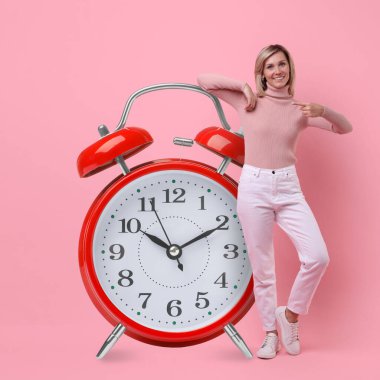 Pembe arka plandaki büyük çalar saati gösteren kadın.
