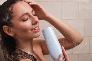 Happy woman with bottle of shampoo in shower at home. Washing hair