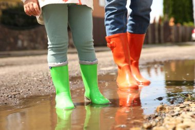 Anne ve kızı lastik botlar giyip açık havada su birikintisinde dikiliyorlar.