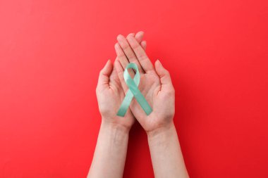 Woman holding turquoise awareness ribbon on red background, top view