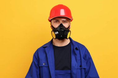 Solunum maskeli ve sarı arka planda kasklı bir adam.