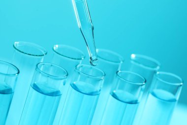 Açık mavi arka planda pipetten test tüpüne sıvı damlatılıyor, yakın plan. Laboratuvar analizi