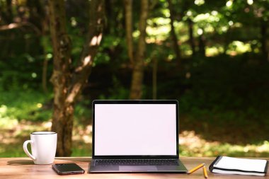 Diz üstü bilgisayar, defter, kalemler, bardak ve akıllı telefon dışarıda, tahta masada. Uzak çalışma