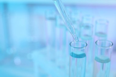 Açık mavi arka planda pipetten test tüpüne sıvı damlatılıyor, yakın plan. Metin için boşluk