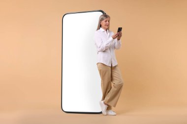 Büyük akıllı telefonun yanında koyu bej arka planda cep telefonu tutan mutlu olgun bir kadın.