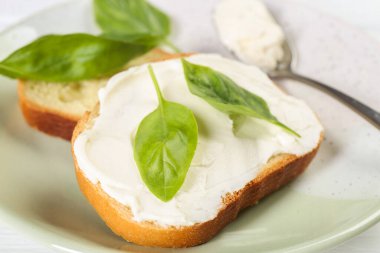 Krem peynirli ekmek parçaları ve beyaz ahşap masada fesleğen yaprakları, yakın plan.