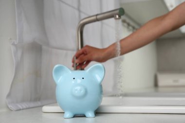 Water saving concept. Woman checking water flow in kitchen, focus on piggy bank clipart