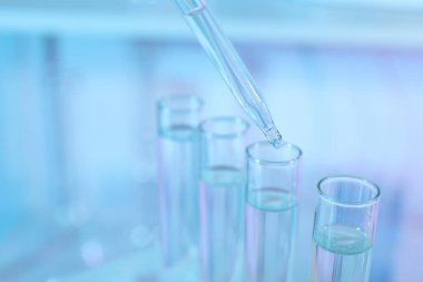Açık mavi arkaplanda pipetten test tüpüne sıvı damlatılıyor, yakın plan.