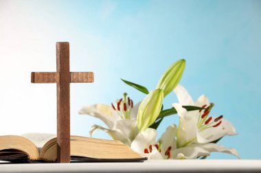 Wooden cross, Bible and beautiful lily flowers on white table against light blue background, closeup. Religion of Christianity clipart