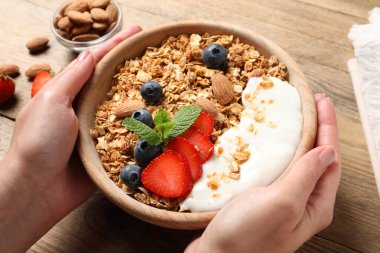 Ahşap masada meyveli, bademli ve yoğurtlu bir kase lezzetli granola tutan kadın.