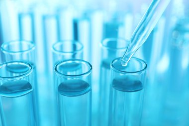 Açık mavi arkaplanda pipetten test tüpüne sıvı damlatılıyor, yakın plan.