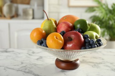 Beyaz mermer masanın üzerinde farklı taze meyveler bulunan cam vazo, yakın plan.