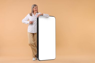 Mutlu olgun kadın koyu bej arka planda büyük bir cep telefonunu işaret ediyor.