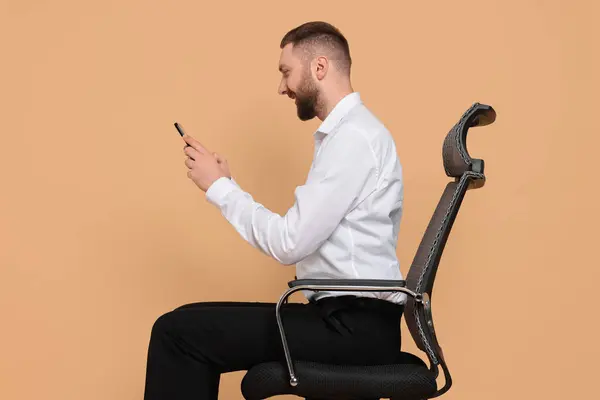 stock image Man with good posture sitting on chair and using smartphone against pale orange background