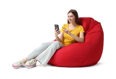 Beyaz arka planda kırmızı fasulye koltukta otururken akıllı telefon kullanan gülümseyen kadın.
