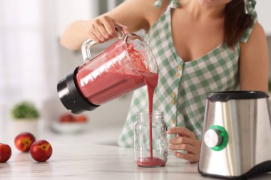 Mutfaktaki beyaz mermer masada, blender bardağından bardağa taze meyve püresi dolduran kadın, yakın plan.