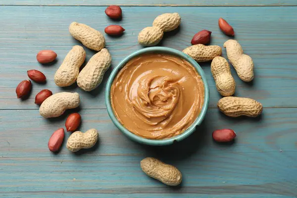 stock image Tasty peanut butter in bowl and groundnuts on light blue wooden table, flat lay